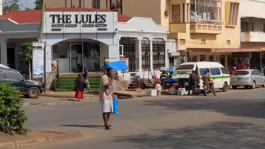 The video depicts a daytime street scene in a bustling urban area. The street is lined with a mix of one- and two-story buildings, some featuring shops and businesses. The storefront "The Lules" is visible on the left side of the frame, advertising underwear, swimwear, and sportswear. Traffic is moderate, with a variety of vehicles including cars, vans, motorcycles, and auto-rickshaws (tuk-tuks) moving along the road. People are seen walking along the sidewalks and crossing the street. One man is on a cellphone holding a blue shopping bag. Several vendors appear to be setting up or packing up items alongside a white van parked near the intersection. The overall environment suggests a typical day in a busy commercial district.
