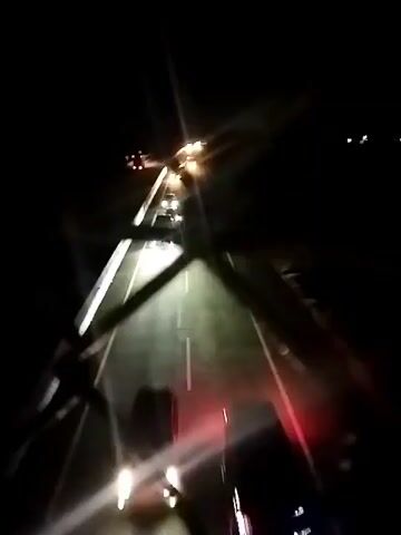 This is a nighttime video of a highway with several cars driving. The camera is positioned above and slightly to the left of the road, possibly from a bridge or overpass, looking down the highway. Headlights from oncoming and passing vehicles illuminate the road. The road appears to be asphalt with white lane markers visible. The sky is dark, indicating it is nighttime. There is a fence or railing in the foreground, out of focus, obscuring the bottom left portion of the frame. The video is shaky and appears to have been recorded on a mobile device.