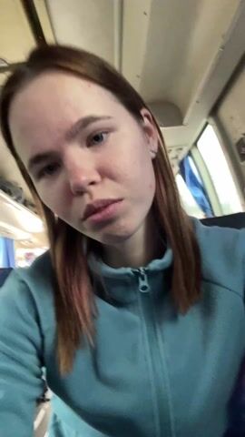 A young woman with fair skin and light brown hair, wearing a light blue fleece jacket, is looking directly at the camera. She is indoors in a vehicle, possibly a bus or train, given the window behind her and the visible interior structure. She is speaking, although the audio is unclear. She appears to be recording a video selfie.