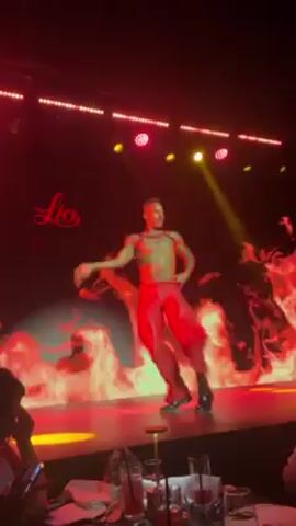 Here is a concise description of the video:

A shirtless dancer with short hair, wearing loose red pants and black shoes, performs on a stage in front of a "Lio" logo and a backdrop of animated flames. The lighting is primarily red, with stage lights overhead. A table with drinks and seated people is visible in the foreground.