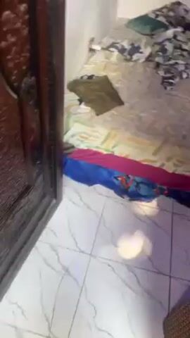 The video begins with a view of a messy bedroom, featuring a bed with disordered bedding and various items scattered on top. The camera then moves outdoors, showing two men standing on a balcony or porch. One man is wearing a yellow shirt, while the other is wearing a maroon hoodie. The man in the hoodie appears to be holding or manipulating something in his hands. The area outside the door seems to be a porch with various objects scattered around.