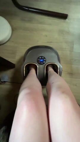 The video shows a person using an electric foot massager. Their feet are inside the device, which has a digital display showing the number "08" and several buttons. The massager is on a wooden floor. The legs of the person using the massager are visible. The audio contains sounds of both the massager and speech in the background.