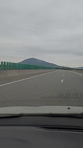The video shows a highway with a mountain in the distance and a concrete barrier with green sound barriers. The camera appears to be mounted inside a vehicle, as the dashboard is visible in the foreground. The road is asphalt with white lane markings. The sky is overcast. The video is 4 seconds long.