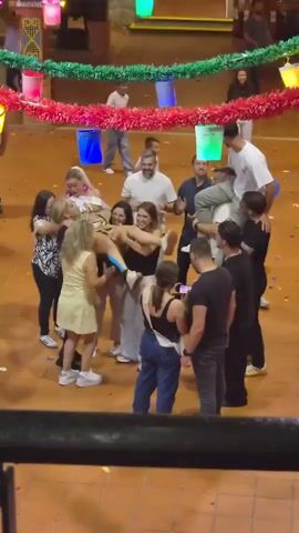 A group of people are gathered in a circle outdoors, under a decorative banner with red and green streamers. Several colorful buckets hang from the banner. In the center of the circle, a woman in a yellow dress is being hugged by other women. One woman is being lifted onto the shoulders of a man. People are smiling and laughing, and there is confetti on the ground. A few children are visible in the background. The ground is paved with reddish-brown tiles.