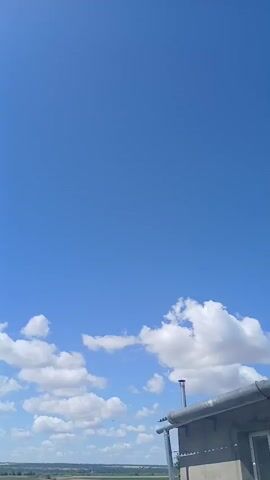 The video shows a clear blue sky with scattered white clouds. In the foreground, there is a roof or ledge, with a pipe extending upwards. Below the sky, a line of trees and some buildings can be seen. The camera pans slightly to the right, revealing more of the landscape.