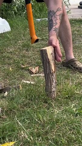 A person is chopping wood with an axe in a grassy area. The person's arms are tattooed. The person is wearing black shorts and croc-like shoes. One piece of wood has already been chopped and lies on the ground.