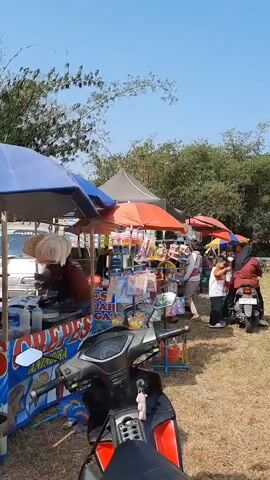 The video shows an outdoor marketplace under a bright blue sky. Several stalls are set up, each with colorful umbrellas providing shade. One stall in the foreground features a motorcycle and a cart selling crepes, with the word "CREPES" prominently displayed in blue and red lettering. Another stall is filled with various colorful items, possibly toys or stationery. People are visible at the stalls, including a vendor handing something to a customer and others browsing or operating their businesses. The ground appears to be covered in dry grass, and trees and bamboo can be seen in the background. The scene suggests a lively and active outdoor market.