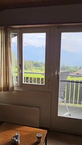 Here is a concise description of the video:

The video shows a view from inside a room, looking out through a set of white-framed windows and a glass door onto a scenic landscape.  The landscape features green fields, trees, and houses nestled in a valley, with snow-capped mountains visible in the distance under a partly cloudy sky. A small balcony is partially visible, with a chair on it. Inside the room, there's a cream-colored curtain on the left, a radiator below the window, and a wooden coffee table with a mug and some small objects on it.