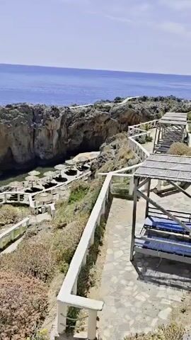 The video shows a view of a rocky coastal area with a small cove-like beach. The beach is nestled between rocky cliffs and features several straw parasols and sun loungers. There are a few people scattered around the beach area, some appearing to be swimming in the water, while others are lounging on the beach. The water in the cove is clear and blue. The landscape is characterized by rugged rocks and sparse vegetation. A white, stone pathway with railings leads down to the beach area from a higher vantage point. The overall impression is of a secluded and scenic beach location.