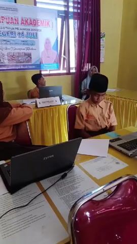 The video shows a classroom setting with students taking a test. There are three students visible in the video, all wearing school uniforms. Each student is sitting at a table with a laptop and papers. The girl on the left is wearing a brown hijab and appears to be looking at her laptop. The boy in the center has his head down, focused on the paper in front of him. The boy on the right is smiling and looking at his laptop screen. 
A banner is hanging on the wall behind the students, which reads "TKA (TES KEMAMPUAN AKADEMIK)" and "UPTD SD NEGERI 14 JULI". 
An adult woman wearing a hijab is also in the room, sitting behind the students.
The room appears to be a classroom, decorated with curtains and a yellow tablecloth on the tables. There are various papers and pens on the tables, suggesting that the students are actively working on their test.
