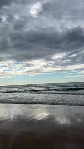 The video begins with a wide shot of a beach, with the ocean meeting the shore, and a cloudy sky overhead. The camera then cuts to a young man with long hair and a mustache, walking toward the camera on the beach. He is wearing a t-shirt, shorts, and a baseball cap. He smiles and gestures with his hand, saying "Good morning." A voice replies "Morning." The camera then cuts back to a wide shot of the beach and ocean.