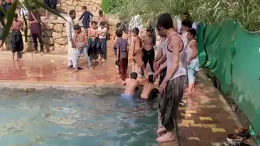A group of young people gather around a rectangular swimming pool, mostly without shirts on. A man wearing a white tank top and baggy pants stands at the edge of the pool. Suddenly, several people jump into the pool. The water splashes, and they swim around. Other people remain on the edge of the pool, observing. The pool area is paved with red and yellow brick-like material. Behind the pool, there is some greenery and a low wall.