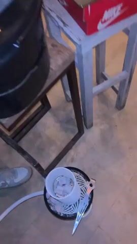 The video shows a person operating a DJ controller, surrounded by hookah equipment. The controller sits atop a black bucket on a small wooden stand. A laptop rests on a Nike shoebox on a nearby stool. Below, a lit hookah bowl with charcoal sits on the floor with a white hose extending from it. The person wears a black jacket, shorts, and gray sneakers. Smoke rises from the hookah and briefly obscures the controller. A stone wall is visible in the background.
