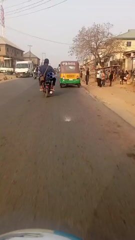 The video shows a street scene in a developing country. The camera is mounted on a moving vehicle, probably a motorcycle or auto-rickshaw, traveling down a paved road. The focus is on a motorcyclist ahead carrying a stack of blue PVC pipes strapped to his back. The road is lined with buildings, some dilapidated and some more modern. There are several auto-rickshaws and other motorcycles on the road. People are walking along the sides of the road. The sky is overcast. There are utility poles and overhead wires visible. The general impression is that of a busy street in a hot, dusty environment.