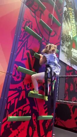 A young girl with blonde hair pulled back in pigtails is climbing a climbing wall indoors. She is wearing a light blue and white striped dress, white leggings, and bright green socks. She is secured by a harness attached to a rope with a clip above her. The climbing wall is brightly colored with a red and black design and has green horizontal handholds. A sign for Jump Yard is visible on the right side of the frame. The girl is actively climbing, moving her hands and feet to ascend the wall.
