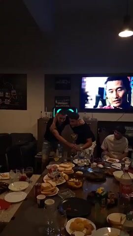 The video shows a room with people gathered around a large table filled with food and drinks. Two men are standing up near the center of the table, holding and waving a gold-colored trophy, as if celebrating. They have their arms around each other and are smiling. A third man sits at the table to the right. A television in the background is displaying a man's face.  Various bottles and glasses are visible on the table. The room is dimly lit, but the lighting becomes brighter as the video progresses.