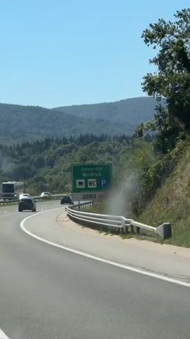 The video shows a view from a moving car on a highway. The highway has multiple lanes and is bordered by a metal guardrail. Green signs indicate upcoming services, including "Odmeriste Modruš," a coffee cup icon, a WC icon, and a parking icon. The signs also state "2000 m." Several cars are visible on the highway, traveling in the same direction. Beyond the highway, rolling green hills covered in trees stretch into the distance under a clear blue sky. The sun is shining brightly, casting shadows. The overall environment is a natural landscape with a man-made highway cutting through it.