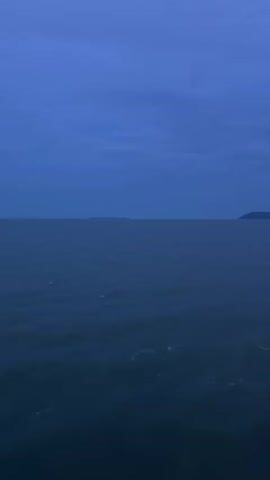 The video begins with a wide shot of a dark blue ocean under a cloudy, twilight sky. In the distance, a faint, dark silhouette of landmasses with mountain-like features is visible. The camera pans slightly to the right, revealing more of the coastline and the vast expanse of water. As the video progresses, the perspective shifts, and the camera appears to be mounted on a moving vessel, likely a boat or ferry, as indicated by the presence of a railing on the right side of the frame. The water shows subtle ripples and occasional white splashes, suggesting movement. The sky remains dark and overcast. The landmasses in the distance become more defined, with some appearing as islands or peninsulas. The overall impression is that of a journey at dusk or dawn across a body of water, with a natural, somewhat subdued landscape in the background.