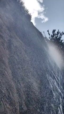 A tall, rocky cliff face is partially obscured by a mist or spray from a waterfall. Water trickles down the rock face in several places, forming darker streaks. Green foliage is visible at the top of the cliff and along the right edge of the frame. The sky above is blue with some white clouds. The camera angle is low, looking up at the cliff.
