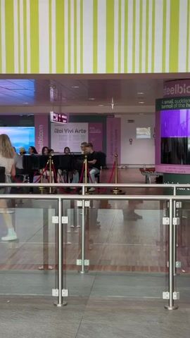 The video shows a group of people gathered around a piano in what appears to be a public indoor space, possibly an airport or a museum. The space is decorated with vertical stripes of green and white on the ceiling and pink and purple signage on the walls. A piano is set up with several people seated at it, some playing, and others watching. A rope barrier separates the viewers from the musicians. People are walking past the scene, with one person in the foreground walking towards the right side of the frame. Another group of young men walks past the piano, one carrying a backpack. A screen displaying colorful graphics is visible in the background. The overall impression is that of a public art installation or performance.