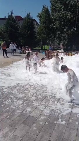 A group of children are playing in a large foam pit on a sunny day. Some children are running through the foam, while others are sitting or playing in it. In the background, there are adults watching and trees.