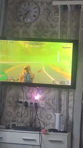 A television screen displays a video game. The game appears to be a third-person shooter set in an outdoor environment with trees and a road. A character wearing camouflage clothing and a backpack is visible on the screen. The scene on the TV screen is bathed in a bright green light. Below the television, several electrical cords are plugged into a power strip, and a white appliance, possibly a kettle, is placed on a white cabinet. A clock with Roman numerals is mounted on the wall above the television.