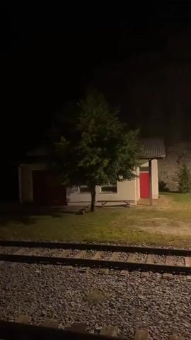 The video shows a nighttime view from a moving train or vehicle alongside a set of train tracks. On the right side of the tracks is a small white building with a red door and a red garage-style door. There is a tree in front of the building, illuminated by a streetlamp. Further down the tracks to the left, another, smaller building is visible. The train tracks are gravel and are in the foreground of the video. The overall scene is dark, with artificial lighting illuminating the buildings and the area around them.