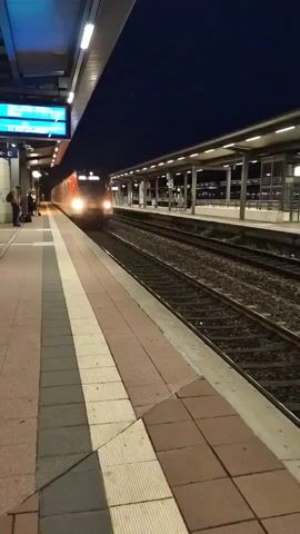A red and white train is approaching a platform at night. The platform is made of concrete tiles and has yellow and white striped markings along the edge. Several people are standing on the platform with luggage, waiting for the train. The train's headlights are on, and its windows are illuminated from within. The platform is covered by a roof supported by columns, and overhead lights illuminate the area. The sky is dark blue, indicating nighttime. A digital display board is visible on the left side of the platform. The train is moving from left to right in the frame and is captured in motion blur, suggesting it is moving at speed.