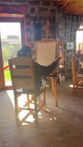 A woman sits at a wooden easel and sketches on a canvas in a sunlit studio. The room is decorated with stone walls, a wooden ceiling, and wooden furniture. Another person sits at a table in the room. A finished stone carving sits on a stool near the table. Natural light floods the room.