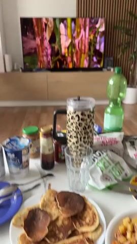 The video shows a living room setting with a large television in the background displaying a nature documentary followed by a scene from a reality show where a woman named Mackenzie is seated. In the foreground, a table is laden with breakfast items. These include a plate of pancakes, a dish of what appears to be hash or potatoes with meat, several containers of yogurt, jars of peanut butter and jam, a leopard-print thermos, a glass, a bottle of green liquid, and various wrappers and utensils. The room has wooden flooring and light-colored walls, with vertical wooden paneling on the wall behind the television. A plant is visible to the right of the television.