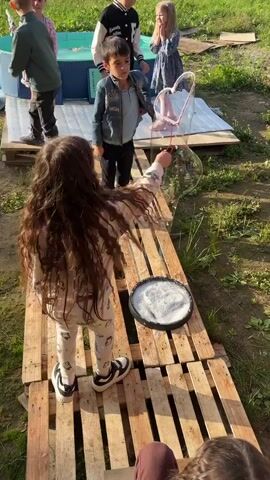 A group of six children are outside in a grassy area. In the foreground, a girl with long curly brown hair is walking on a wooden path made of pallets. She is holding a bubble wand. A round black container filled with soapy water is on the wooden path in front of her. Behind her, two boys and three girls are standing on a raised platform near a blue inflatable pool. The children appear to be engaged in an activity together. The environment is sunny and appears to be a backyard or outdoor play area.