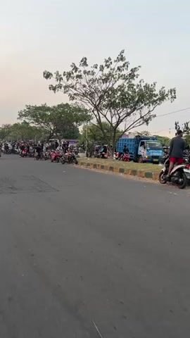 The video shows a street scene with many motorcycles and people gathered. The time is likely late afternoon or early evening, as indicated by the soft, warm lighting and the sky. The street appears to be a road with a paved surface and a curb. On the right side of the road, there is a grassy area with trees and a blue truck. A significant number of motorcycles are parked or being ridden by people. Some individuals are on the motorcycles, while others are standing around. The atmosphere seems casual and social, possibly a gathering or event. The people are dressed in everyday clothing. Some motorcycles are being revved or ridden slowly, suggesting a leisurely or informal activity. The presence of a truck and roadside vegetation indicates an outdoor setting, potentially on the outskirts of a town or in a less urbanized area.