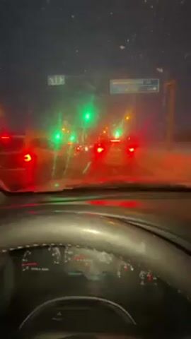 The video shows a driver's point of view in a car stopped at a traffic light during a snowy night. The windshield is wet and has snow on it, obscuring the view slightly. There are cars ahead, their brake lights visible. Street signs are visible in the distance, but the details are difficult to discern. The dashboard of the car is visible, with the steering wheel in the foreground. The speedometer reads zero, indicating the car is not moving. The lighting is poor due to the night and snow. The general atmosphere is that of a cold and snowy winter night. The road has some visible snow covering. The sound of music is playing in the background.