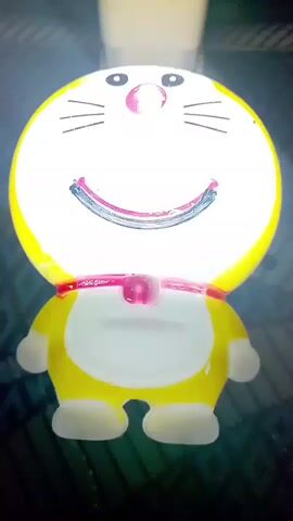 The video displays a yellow, cartoonish robot figurine resembling Doraemon. The figurine is lit from within, making its face appear bright and illuminated. Its eyes are black dots, and it has two black whiskers on each side of its head. A pink, bell-shaped object is visible around its neck. The figurine is standing upright, facing the camera. The background is dark and appears to be indoors, with some patterned texture visible on the surface it is resting on.