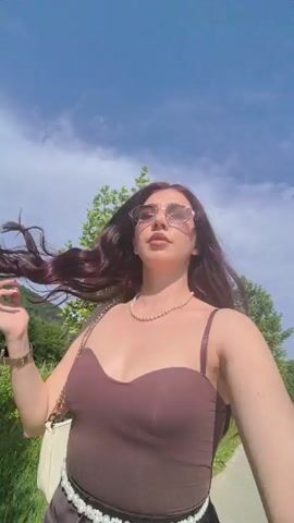 A woman with long, dark, reddish-brown hair, wearing sunglasses, a brown top, and a pearl necklace, walks along a paved path. She holds the camera in front of her, facing herself, as she walks. The camera is held low and points upwards, showing her from the waist up. Lush green foliage and trees line the path on both sides. In the background, there is a vineyard and hills under a clear blue sky. The woman smiles and at one point adjusts her hair.