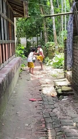 Two young boys are playing in a narrow outdoor passageway. The path is made of weathered bricks and dirt, flanked by a building on one side with a wooden frame and another brick structure with a hanging cloth on the other. The boys are wearing light-colored shirts and yellow pants. They are facing each other, then one leans over, and the other playfully jumps on his back, grabbing around his neck. The area is surrounded by lush green vegetation, including trees and leafy plants. The overall impression is a simple, rustic scene of children playing in a rural environment.