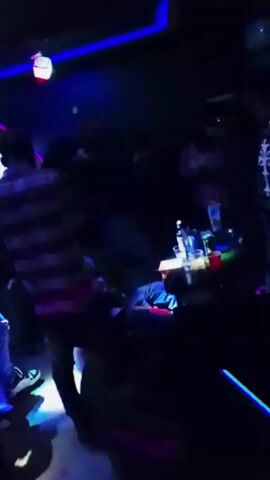 The video shows a dimly lit interior space with a number of people standing and moving around. There are colorful lights, including green, blue, and red, that illuminate the scene and create a party-like atmosphere. Several red plastic cups and what appears to be a bottle of liquor are visible in the foreground. The silhouettes of individuals are seen in various positions, but their features are indistinct due to the low lighting. The overall environment suggests a social gathering or party, possibly at a nightclub.