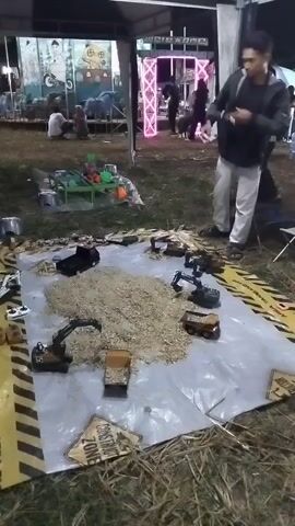 The video shows an outdoor event at night. In the foreground, there is a tarp laid out on the grass with several toy construction vehicles arranged around a pile of wood shavings or mulch. There are also remote controls on the tarp, suggesting the toys are radio-controlled. In the background, there are people, tents, and bright lights, including a pink light installation. Some people are gathered around what appears to be a stage or display area. The overall atmosphere seems to be that of a casual outdoor fair or market.