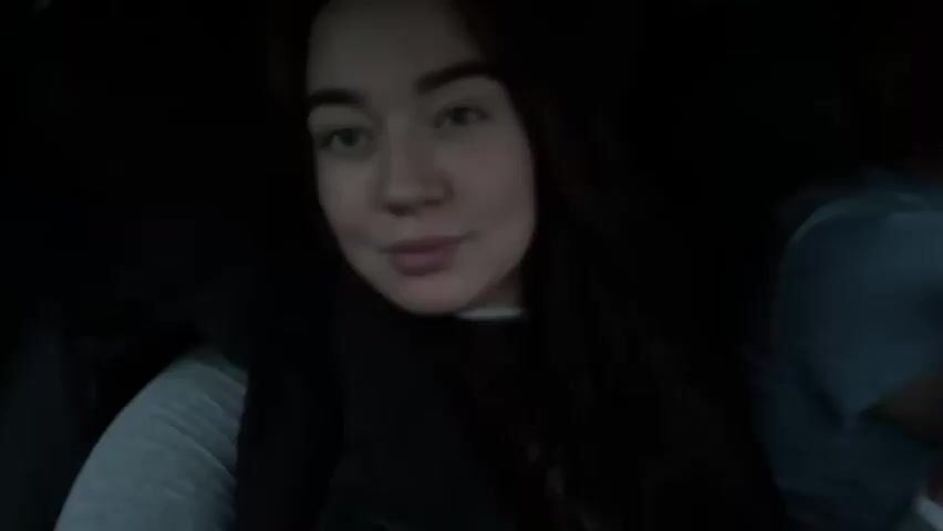 The video shows a close-up of a young woman with long, dark hair and blue eyes. She is looking at the camera and smiling slightly. She is wearing a grey long-sleeved shirt and a dark vest. Her face is well-lit, but the rest of the scene is dark. There is another person partially visible on the right side of the frame, wearing a grey shirt. The woman appears to be inside a car.
