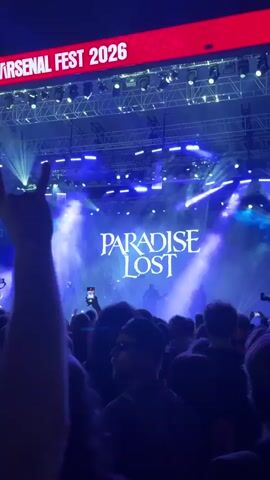 The video displays a concert crowd with a stage in the background. The stage features a large screen displaying the words PARADISE LOST in white lettering, illuminated by blue and white stage lights and smoke effects. A banner above the stage reads ARSENAL FEST 2026. The crowd is densely packed, with many individuals holding up their phones to record the performance. The lighting is dim, creating a dramatic atmosphere. The audience members are mostly facing the stage, engrossed in the event.