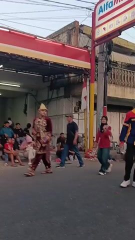 The video shows a street scene with several people. In the foreground on the left, a young person in traditional clothing stands with a child in an orange shirt. To the right, a person in a red, yellow, and blue jacket walks away from the camera. In the background, there is a store with a red and white sign that says "Alfamart". Other people are visible standing and sitting around the street. There are also several motorcycles and carts. The sky is overcast, suggesting it may be morning or evening.