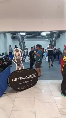 A group of people, mostly men and boys, are gathered in an indoor space. There are cardboard cutouts of anime characters, likely from the Beyblade franchise, present in the scene. A large pop-up banner with the Beyblade X logo is visible on the floor. Some individuals appear to be interacting with each other, possibly participating in a competition or event related to Beyblade. The environment suggests an event space or a promotional area with banners and displays. The lighting appears to be artificial.
