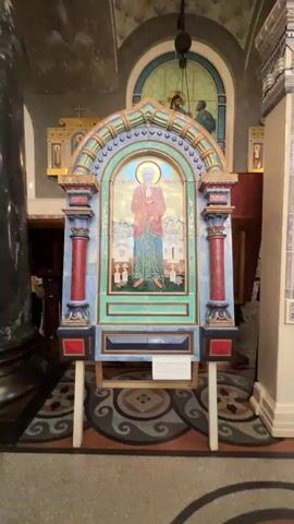 The video shows two tiled iconostases inside what appears to be a church. The first iconostasis, on the left, features a mosaic of a person in a pink robe and green skirt, standing in front of buildings. It is supported by a wooden easel. The second iconostasis, on the right, is larger and more ornate, with blue and white tiles. It displays a mosaic of a man in a black coat and breeches, holding a scroll. Behind the iconostases, a stained-glass window depicting two figures can be seen. The floor has a patterned carpet. The lighting is dim, with a chandelier visible in the background.