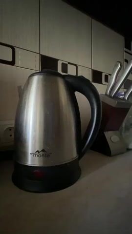 The video begins with a close-up shot of a stainless steel electric kettle with a black handle and base. A red indicator light is visible on the front of the base. The kettle is positioned on a countertop, with a backsplash of tiled squares in the background. To the right of the kettle, a wooden knife block holding several knives is visible. In the first second of the video, a hand enters the frame from the right and grips the handle of the kettle. In the second second, the hand presses down on the handle, and the red indicator light on the kettle's base illuminates. The hand remains on the handle for the remainder of the video, and the kettle sits on the counter with the light on. The environment appears to be a kitchen.