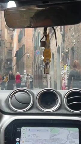 The video shows a car driving down a narrow street in Siena, Italy. The street is crowded with pedestrians walking in both directions. The buildings on either side of the street are made of brick and have arched windows and doorways. The car has a rearview mirror with a hanging air freshener that says "Arbre Magique." The dashboard has three circular air vents and a navigation system displaying a map of Siena. The car is moving slowly.