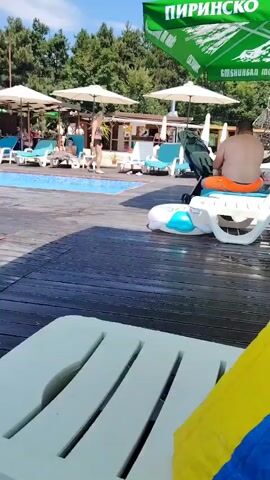 The video opens with a poolside scene. In the foreground, a white lounge chair with yellow and blue towels is visible. The camera then pans to the left, revealing a swimming pool with blue water and a wooden deck. Several lounge chairs and umbrellas are set up around the pool. A few people are relaxing on the lounge chairs, and some are in the pool. A young boy is near the edge of the pool, partially submerged in the water. In the background, trees and a fence are visible. A large green umbrella with white lettering is positioned at the top of the frame. The overall atmosphere appears to be that of a sunny day at a resort or public pool.