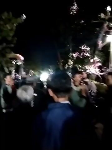A crowd of people are gathered outdoors at night, illuminated by artificial lights. The scene appears to be a public gathering or event. Some individuals are wearing hats and jackets. Trees and foliage are visible in the background. The footage is shaky and appears to be recorded on a mobile device. There are lights and some structures that could be part of a stage or performance setup. In some frames, there are blurred figures and objects moving quickly, suggesting movement within the crowd or by the camera operator. There is no clear indication of any specific activity beyond the gathering itself.