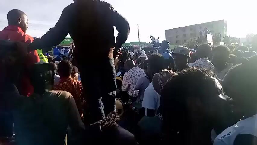 A large crowd of people is gathered outdoors on a very bright and sunny day. The scene appears to be a public event, possibly a rally or community gathering, in an urban or semi-urban setting, with buildings and power lines visible in the background.

Several individuals are elevated, standing on what looks like the back of a truck or a makeshift platform, towering over the main crowd. Two men in particular, one in a red shirt and another in a dark hoodie, are prominent on this platform. The crowd below them is dense, with people closely packed together, all generally facing in the same direction as those on the platform.

Many people in the crowd, including some on the elevated platform, are holding up smartphones, seemingly recording the event or taking pictures. The bright sunlight creates some glare in the footage.

Visible details include:
- A white sports jersey with "ILLS 44" printed on it.
- A military-style shirt with "MINO" written on the back.
- A red shirt with partial text "VIRI" visible.
- Some motorbikes are partially visible at the bottom of the frame, possibly used for transport or as part of the crowd.

The atmosphere appears to be active and engaged, with people looking intently and listening. The audio includes voices speaking, suggesting a public address or speech is taking place.