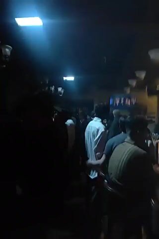 The video shows the interior of a bar. In the first segment, patrons are gathered around a bar area, with a television screen visible in the background displaying what appears to be a sporting event. The lighting is dim and warm. The second segment introduces blue lighting, possibly from a DJ booth or stage, creating a more club-like atmosphere. Patrons are still present, and some are interacting near the bar. The third and fourth segments return to a warmer, more subdued lighting. The camera pans across the bar area, showing shelves stocked with bottles of alcohol behind the bar, and more patrons mingling. A couple is visible interacting at the bar in one shot. The final segment is very dark, with only faint lights visible, suggesting the end of the recording or a significant decrease in ambient light. Throughout the video, the environment appears to be a busy social setting.