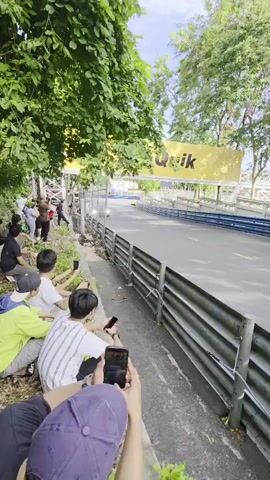 A group of people are watching race cars on a track. The cars are going by quickly, and the people are taking pictures and videos with their phones. There are trees and a fence along the side of the track. A large yellow banner with the word "Quik" is visible in the background.